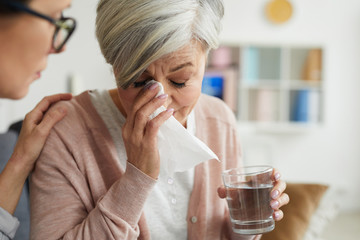 Close up portrait of elegant senior woman crying during therapy session and holding glass of water with female psychologist comforting her