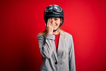 Young beautiful blonde motorcyclist woman wearing motorcycle helmet over red background covering one eye with hand, confident smile on face and surprise emotion.