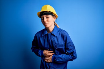 Young beautiful worker woman with blue eyes wearing security helmet and uniform with hand on stomach because nausea, painful disease feeling unwell. Ache concept.