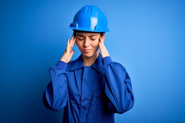 Young beautiful worker woman with blue eyes wearing security helmet and uniform suffering from headache desperate and stressed because pain and migraine. Hands on head.