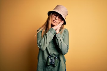 Fototapeta premium Beautiful blonde explorer woman with blue eyes wearing hat and glasses using binoculars sleeping tired dreaming and posing with hands together while smiling with closed eyes.