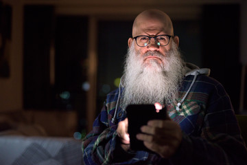 Mature bald bearded hipster man using phone at home late at night in the dark