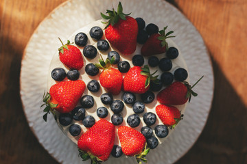 Top view of beautiful delicious summer cake decorated with strawberries and blueberries on a plate on wooden table background
