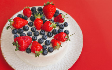 Appetizing cake with white whipped cream frosting decorated with blueberries and strawberries on beautiful plate on red background with copy space. Home made dessert