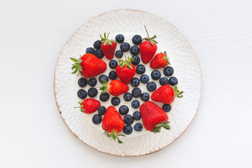 Top view of white cream cake or pie decorated with strawberries and blueberries on white handmade plate on white wooden table background. Summer dessert