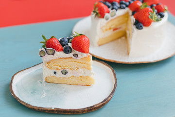 A piece of sponge cake with white whipped cream decorated with strawberries and blueberries on beautiful handmade dishes. Delicious dessert with summer berries on blue and red background