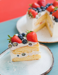 A piece of celebrative sponge cake with white whipped cream decorated with strawberry and blueberry on beautiful handmade plates. Delicious dessert with summer berries on blue and red background
