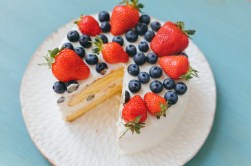 Delicious celebrative home made cut sponge cake with cheese cream on white plate on light blue wooden background. A dessert is decorated with fresh blueberries and strawberries. Selective focus