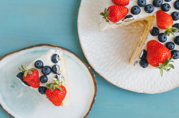 Top view of delicious sweet summer home made cut cake on interesting handmade dishes on light blue wooden background. A dessert is decorated with fresh blueberries and strawberries