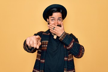 Young handsome hispanic bohemian man wearing hippie style and boho hat laughing at you, pointing finger to the camera with hand over mouth, shame expression
