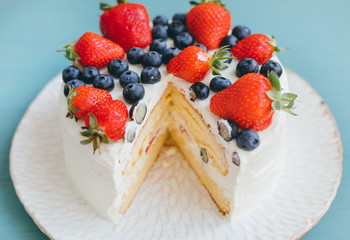 Beautiful delicious cut sponge cake with whipped cream and fresh summer berries on handmade plate on light blue background. Dessert with blueberries and strawberries. Selective focus