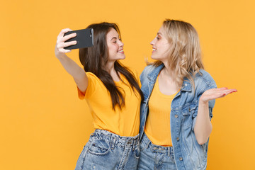 Two smiling young women girls friends in casual t-shirts denim clothes posing isolated on yellow background studio. People lifestyle concept. Doing selfie shot on mobile phone looking at each other.