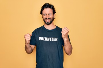 Young handsome hispanic volunteer man wearing volunteering t-shirt as social care very happy and excited doing winner gesture with arms raised, smiling and screaming for success. Celebration concept.