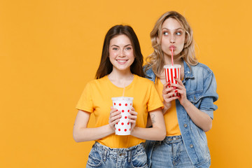 Two smiling young women girls friends in casual t-shirts denim clothes isolated on yellow background studio portrait. People lifestyle concept. Mock up copy space. Hold plastic cups of soda or cola.
