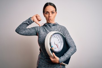 Young fitness woman wearing sport workout clothes holding scale for healthy weight with angry face,...