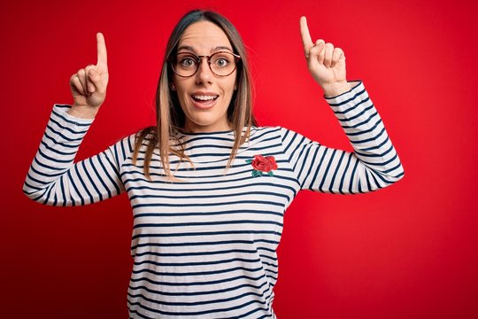 Young beautiful blonde woman with blue eyes wearing glasses standing over red background smiling amazed and surprised and pointing up with fingers and raised arms.