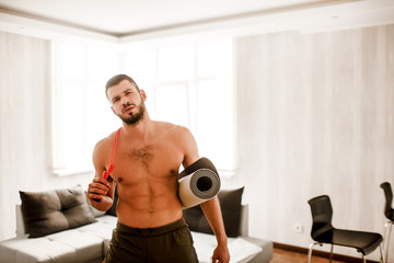 Fototapeta na wymiar Young man doing exercising at home on the couch. He is holding a yoga mat and a skipping rope and look at the camera