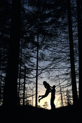 Fototapeta premium From below view of silhouettes of loving couple posing in evening forest. Wild nature with amazing views, summer sunset, man holding woman while jumping and feeling happy. Nature, love concept.