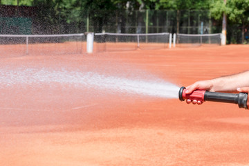 Hose sprinkler in the hand of a worker, jet and spray of water and blurry clay tennis courts background. Maintenance of clay tennis court. Service and preparation of the court. Close up. Copy space.