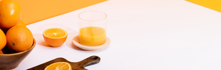 fresh orange juice in glass near ripe oranges in bowl and wooden cutting board on white surface on orange background, panoramic shot