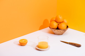 fresh orange juice in glass near ripe oranges in bowl and knife on white surface on orange background