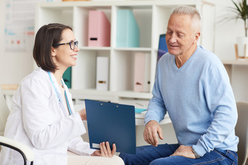 Portrait of smiling female doctor showing clipboard to senior patient during consultation in clinic