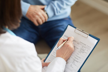 High angle close up of female doctor holding clipboard and filling patients card while consulting unrecognizable senior patient in clinic, copy space