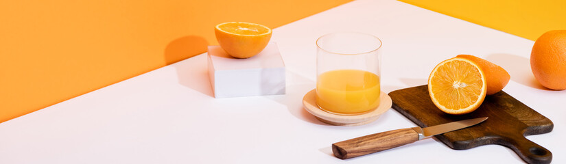 fresh orange juice in glass near ripe oranges, wooden cutting board with knife on white surface on orange background, panoramic shot