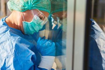 Mediziner mit Schutzkleidung bei Coronavirus Pandemie lehnt ermüdet Kopf an Fenster