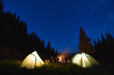 Back view, tourists sitting by brightly blazing campfire between tents against the backdrop of...