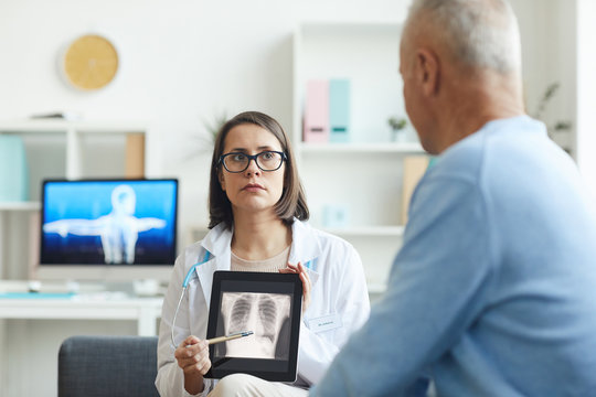 Portrait of serious female doctor holding x-ray image of lungs and chest while consulting senior patient in clinic, copy space