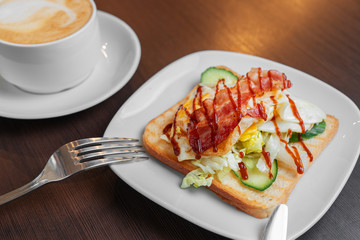 An egg and bacon sandwich on a white plate next to a cappuccino on a dark wooden table.