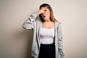 Young beautiful brunette sportswoman wearing sportswoman training over white background peeking in shock covering face and eyes with hand, looking through fingers with embarrassed expression.