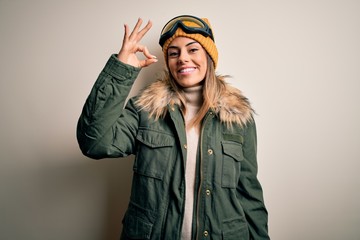 Young brunette skier woman wearing snow clothes and ski goggles over white background smiling positive doing ok sign with hand and fingers. Successful expression.