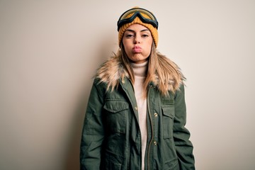 Young brunette skier woman wearing snow clothes and ski goggles over white background puffing cheeks with funny face. Mouth inflated with air, crazy expression.