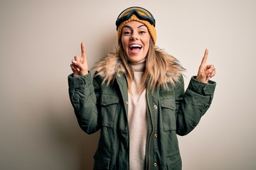Young brunette skier woman wearing snow clothes and ski goggles over white background smiling amazed and surprised and pointing up with fingers and raised arms.