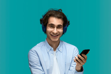 Funny message. Portrait of a young beautiful man wearing white t-shirt and blue shirt in blue headphones holding mobile phone in hand and looking at camera with laugh