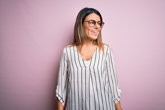 Young beautiful woman wearing casual striped t-shirt and glasses over pink background looking away to side with smile on face, natural expression. Laughing confident.