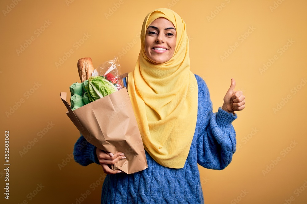 Sticker young beautiful brunette arab woman wearing islamic hijab holding paper bag with food happy with big