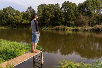 Fototapeta na wymiar A young guy in denim shorts and a longsleeve stands on a wooden structure by the river, with his hands in his pockets and looking at the river, back and side view.