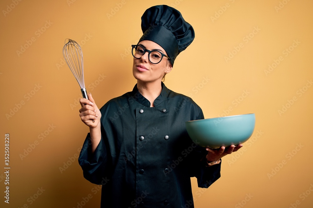 Poster Young beautiful brunette chef woman wearing cooker uniform and hat holding bowl and whisk with a confident expression on smart face thinking serious