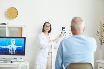 Portrait of young female ophthalmologist pointing at eye chart while testing eyesight of senior patient, copy space