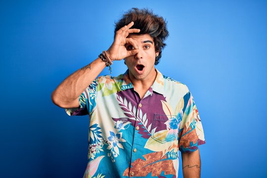 Young handsome man on vacation wearing summer shirt over isolated blue background doing ok gesture shocked with surprised face, eye looking through fingers. Unbelieving expression.