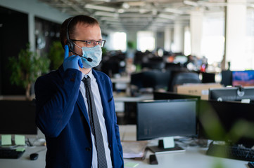 A man in latex gloves and a medical mask is talking to a client over the headset. A male office manager answers calls. Work during the coronavirus. White-collar tie and jacket.