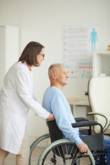 Vertical side view portrait of smiling female doctor pushing senior man in wheelchair for examination or consultation in clinic, copy space