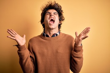Young handsome man wearing casual shirt and sweater over isolated yellow background crazy and mad shouting and yelling with aggressive expression and arms raised. Frustration concept.