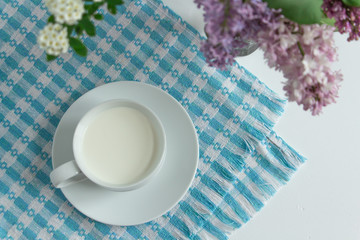 a white cup of fresh milk on a white background with sprigs of white and purple flowers that complement the composition, tasty, healthy, atmospheric
