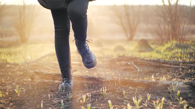 Cinematic shot in front of young girl strolling in trendy leggings and stylish black high-soled boots on ash ground after fire against a field under sunset. Person steps on burned grass in the spring.