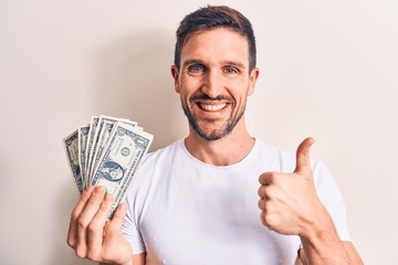 Young handsome man holding bunch of dollars banknotes over isolated white background smiling happy and positive, thumb up doing excellent and approval sign