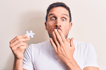 Young handsome man holding piece of puzzle standing over isolated white background covering mouth...